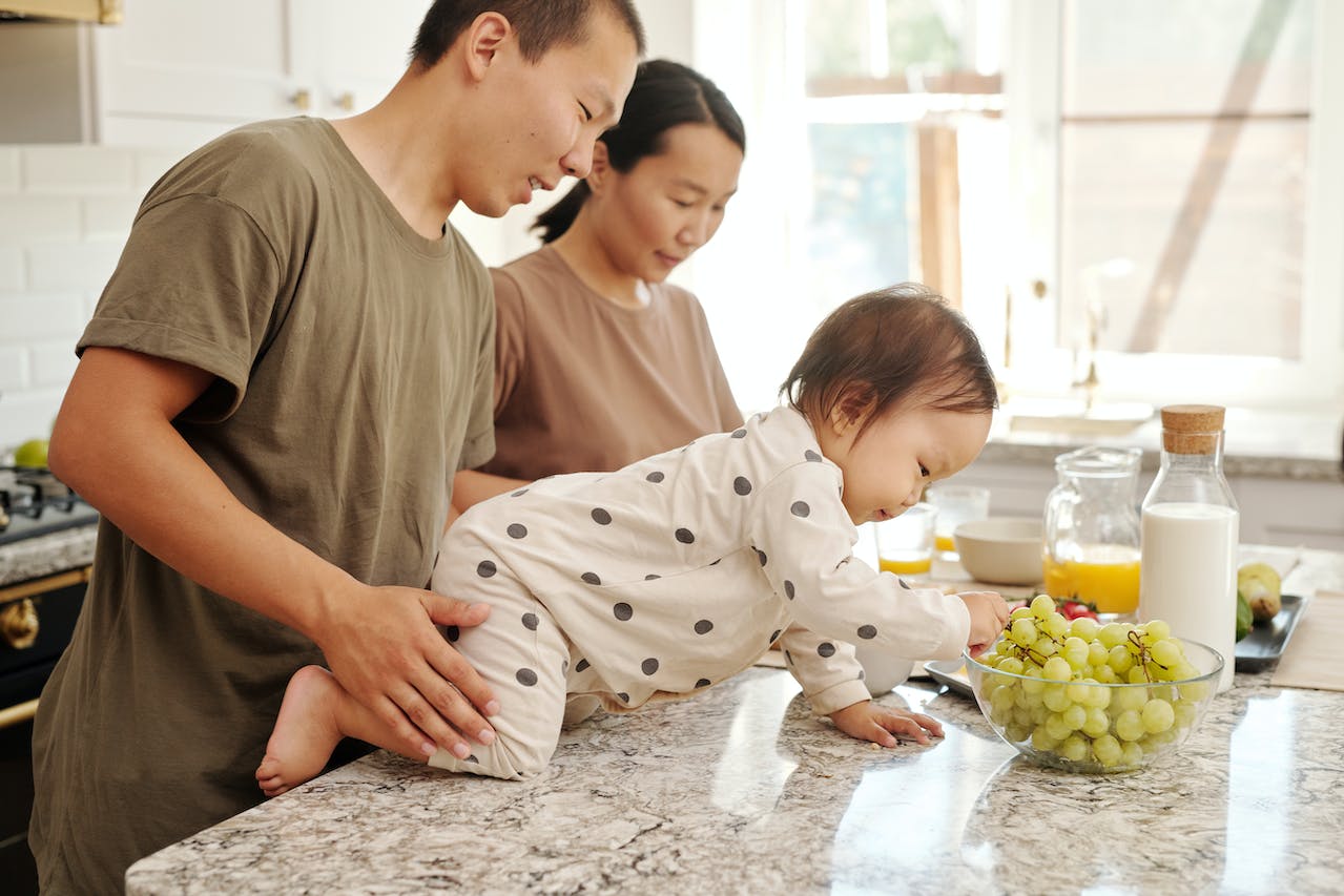 A father holding a baby that's crawling on the counter towards grapes and the mother cooking next to them,