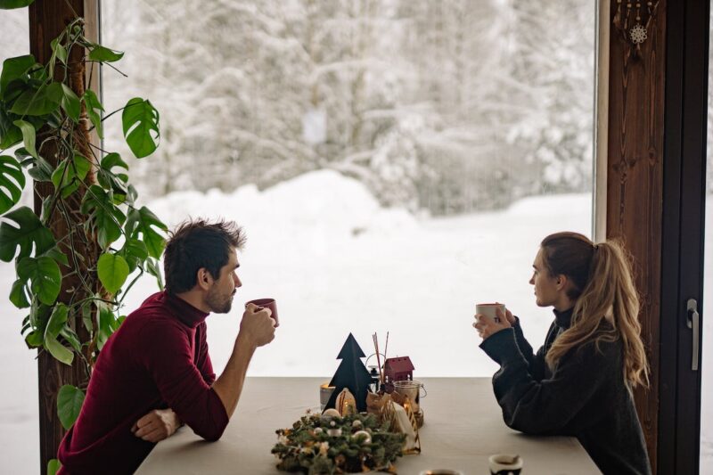 A couple in front of a window in their dining room enjoying warm cups of coffee looking out to the snowy landscape.